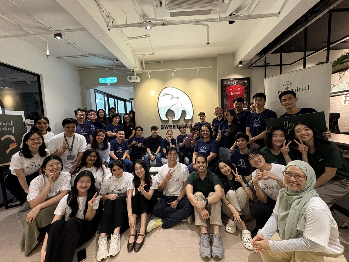 A large group of smiling colleagues posing together in a bright modern office space. Many are wearing matching dark blue t-shirts with a circular white logo, while others wear casual white and green tops.